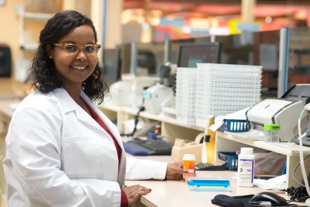 A pharmacy student in a white coat and smiling at the camera stands behind a counter in a pharmacy