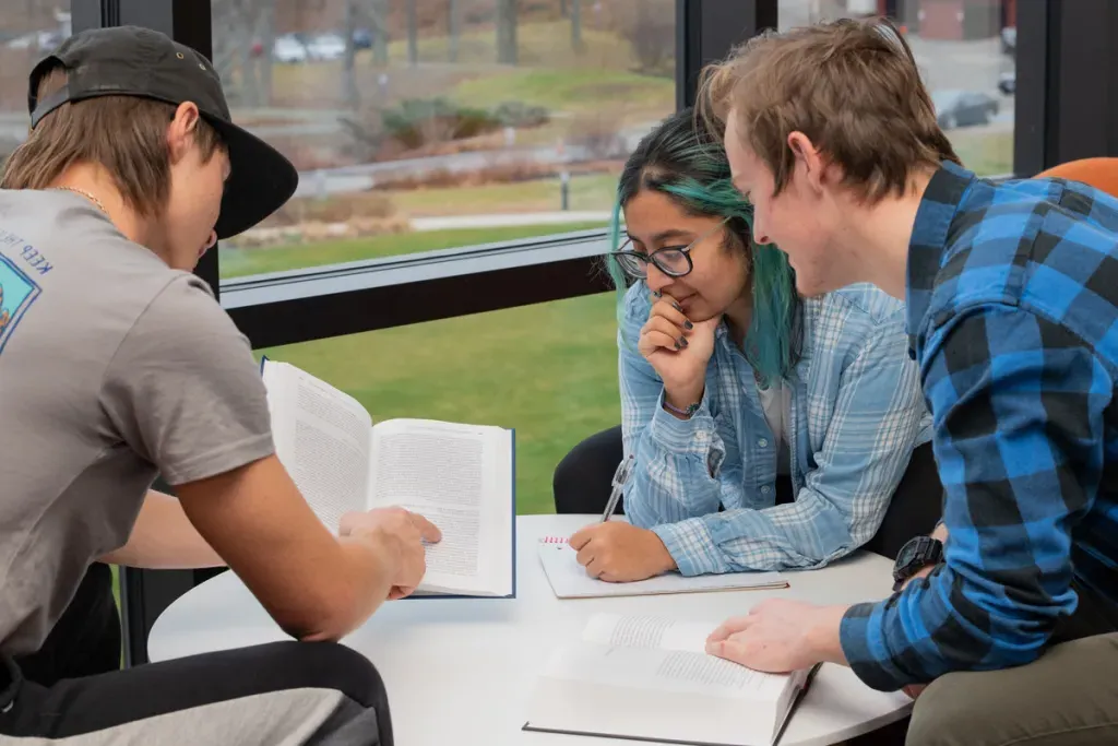 Three students sit around a table with open books scattered across the top