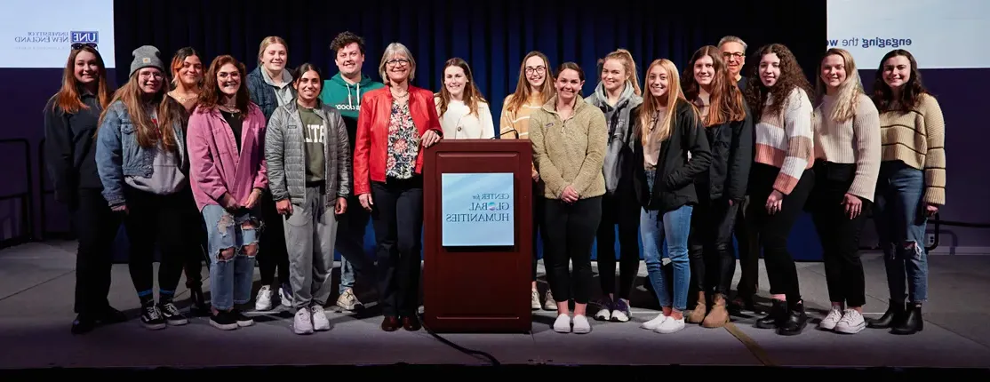 A large group of students stand on stage with a C G H lecturer